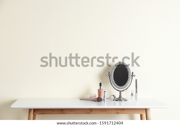 Mirror and makeup products on white table near light wall. Space for text