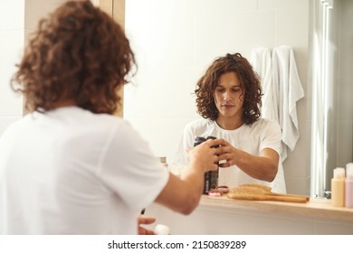 Mirror Reflection And Back View Of Young European Man Taking Shaving Foam From Under Mirror Shelf In Bathroom. Handsome Curly Guy Wearing T-shirt. Modern Domestic Lifestyle. Hygiene And Face Skin Care