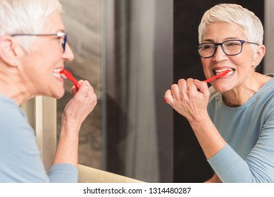 Mirror reflection of beautiful mature woman brushing her teeth with red toothbrush, Daily dental hygiene routine concept - Powered by Shutterstock