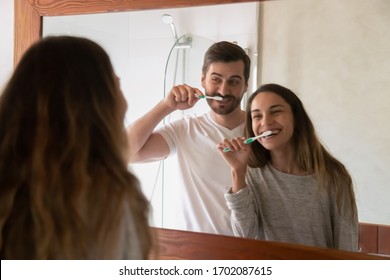 Mirror reflection childish young family couple having fun while cleaning teeth together in modern bathroom. Joyful smiling positive millennial spouses joking doing morning oral hygiene routine. - Powered by Shutterstock