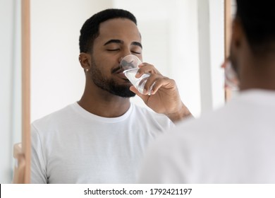 Mirror reflection close up satisfied African American young man wearing white t-shirt drinking pure clean mineral water, standing in bathroom with closed eyes, enjoying morning healthy habit - Powered by Shutterstock