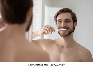 Mirror Reflection Happy Young 30s Bare European Guy Brushing Teeth With Tooth Brush And Toothpaste, Enjoying Oral Dental Morning Cleaning Routine In Bathroom, Involved In Whitening Teeth Procedure.