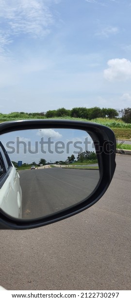 The mirror or side\
mirror of a white car, where you can see the view of a smooth road\
with trees around it.