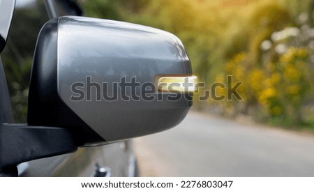 Mirror wing of gray with turn light signal. Blurred of asphalt road and trees with yellow flower for background.