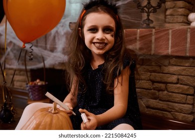 Mischievous child  little girl dressed as witch and spider painted her face  smiles looking at camera  drawing scary face pumpkin  Jack  O  Lantern  Concept happy Halloween party
