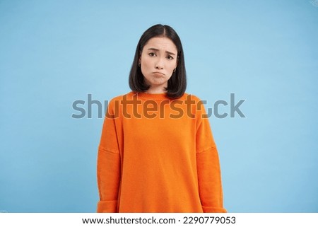 Miserable asian girl, sulks and looks sad, frowns and pouts disappointed, feels like loser, stands over blue background.