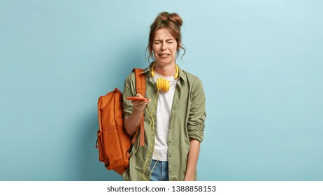 Miserable dejected hipster girl cries and sobs from despair, holds modern smart phone, has problem, cannot browse music app, dressed in casual shirt and jeans, carries rucksack on one shoulder - Powered by Shutterstock