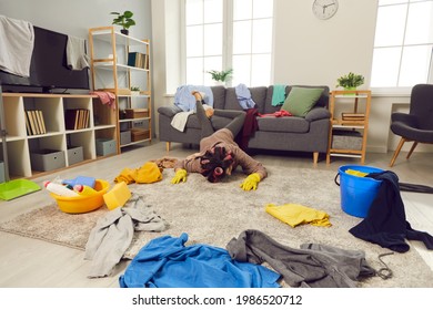 Miserable Tired Housewife Fallen On Floor While Cleaning Up Crazy Mess In Her House. Exhausted Young Woman Lying Face Down On Floor In Living-room Amidst Chaos Of Scattered Untidy Clothes