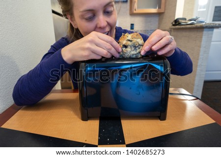 Misguided woman attempts to stuff a large blueberry scone into a small toaster slot, meant for bread
