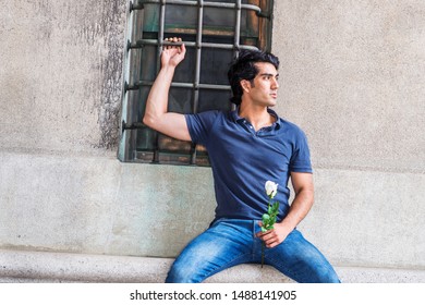 I missing you and thinking of you. Young Man wearing blue short sleeve shirt, jeans, sitting against wall with window on street in New York City, holding white rose flower, seriously looking away.
