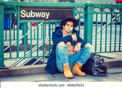 I Missing You. Wearing Jacket With Hood, Jeans, Boot Shoes, Fedora Hat, Leather Bag On Ground, A Guy With Freckle Face, Sitting On Street By Subway Sign In New York, Smelling White Rose, Thinking.
