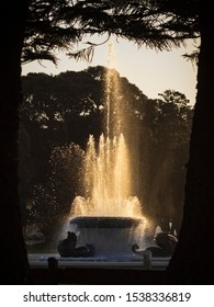 Mission Bay Fountain Framed By Silhouette Trees And Glowing On A Golden Hour Dawn Sunrise Morning In A Public Garden Park In Auckland City New Zealand