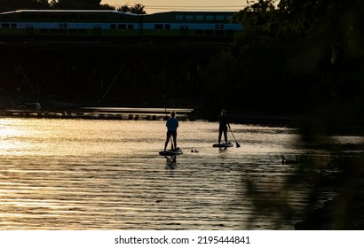 Mississauga, Ontario - August 28, 2022: Silhouette Of A Couple Of Women On Stand Up Paddle Board Going Up Credit River And A GO Train Is Passing By The Bridge Above