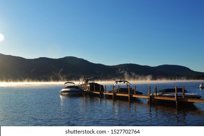 Mist Floats Above Lake George In New York In The Morning With The Blue  Sky Overhead, Mountains Rising In The Distance And Boats Resting At The Dock.