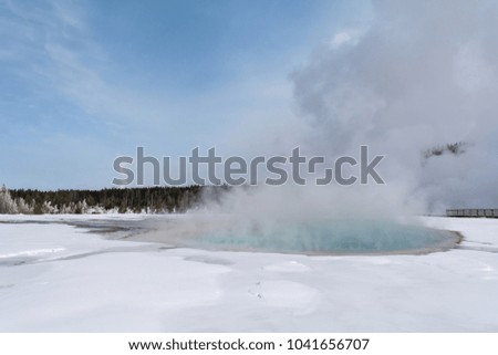 Mist rises from the azure water of a geyser pool sitting in a snowy field.
