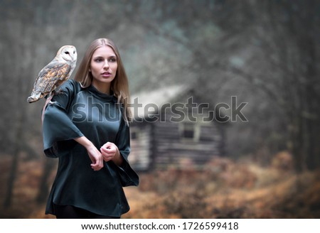 Misterioius young blond girl with a barn owl white bird in a foggy forest and an old hut background in a blue dress