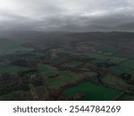 Misty aerial view of lush green fields, patchy woodlands, and distant mountains under an overcast sky, creating a serene and moody landscape.