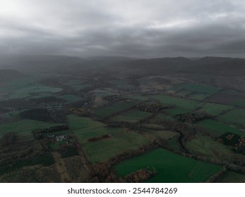 Misty aerial view of lush green fields, patchy woodlands, and distant mountains under an overcast sky, creating a serene and moody landscape. - Powered by Shutterstock