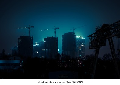 Misty cityscape at night time. Cranes over houses under construction.