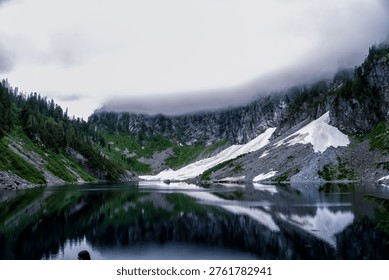 Misty clouds hover above jagged mountain peaks. Snow patches cling to steep, rocky slopes. Deep lake mirrors the sky and mountains perfectly. Trees cling to cliffside, green and resilient