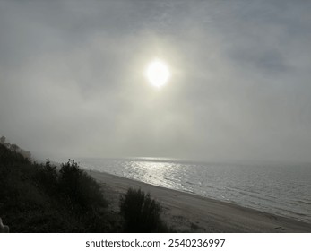 A misty coastal landscape with the sun shining through a hazy sky, illuminating the beach and casting a soft glow over lush greenery on the cliffs, creating a serene, atmospheric scene. - Powered by Shutterstock