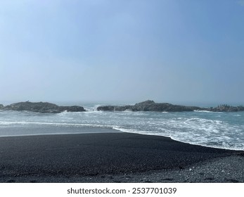 Misty coastal view of waves washing over a black sand beach with rocky outcrops in the distance - Powered by Shutterstock