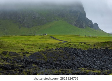 Misty dramatic landscape featuring lush green mountain slopes with dark basalt lava rocks in foreground and tourists exploring pristine