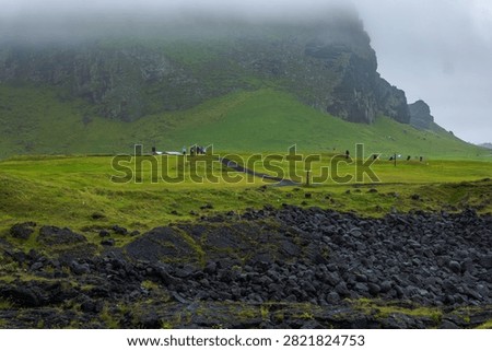 Misty dramatic landscape featuring lush green mountain slopes with dark basalt lava rocks in foreground and tourists exploring pristine