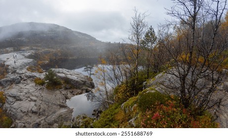 A misty landscape with rocky terrain, sparse trees, and a calm lake reflecting the overcast sky. The scene is serene and natural, with autumn foliage adding color to the rugged environment. - Powered by Shutterstock