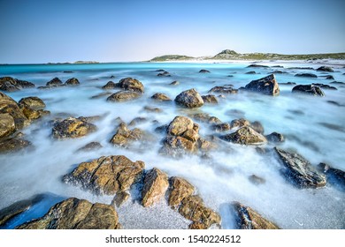 A misty long exposure of rocks on the shoreline. Blues waters. The beach continues in the background. 