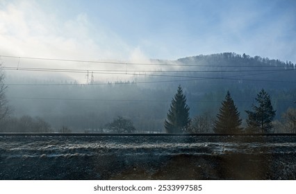 A misty morning in the mountains with a railway track in the foreground. Power lines stretch across the frame as sunlight filters through the fog, casting soft light on the forested hillside. - Powered by Shutterstock