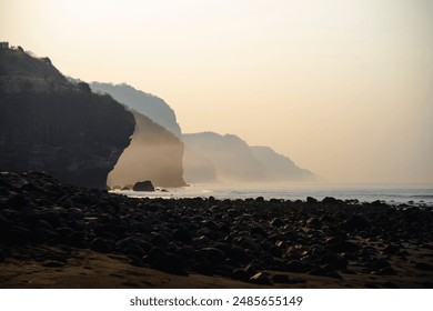 Misty morning at a secluded beach with rocky terrain and distant cliffs enveloped in fog. The sunrise casts a golden hue over the serene landscape, natural beauty and tranquility of the scene - Powered by Shutterstock