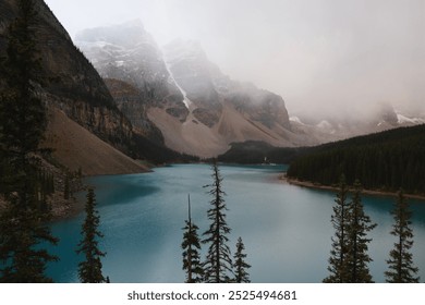 A misty mountain landscape frames a turquoise lake. Snow-capped peaks rise majestically above a dense forest, reflecting in the calm waters. The scene is peaceful and serene. - Powered by Shutterstock