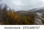 A misty mountain landscape with rocky terrain and autumn foliage. The scene features trees with yellow leaves, rocky outcrops, and a cloudy sky, creating a serene and atmospheric environment.