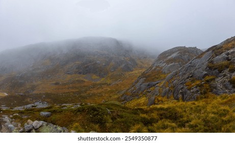 A misty mountain landscape with rocky terrain and sparse vegetation, featuring a cloudy sky obscuring the mountain peaks. - Powered by Shutterstock