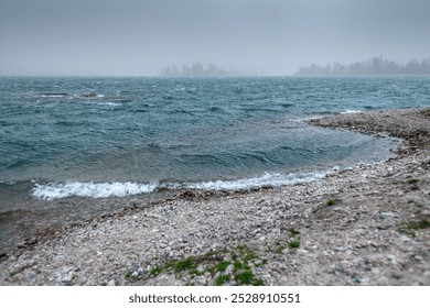 a misty and overcast scene of a river or lake during a storm. The water is rough, with small waves crashing against the rocky shore. The horizon is obscured by fog or mist, giving the distant trees an - Powered by Shutterstock