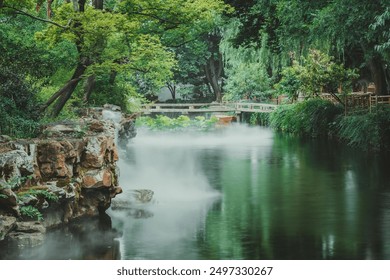 A misty pond with a stone bank and a wooden bridge. Lush greenery and foliage surround the water, creating a serene and tranquil atmosphere. - Powered by Shutterstock