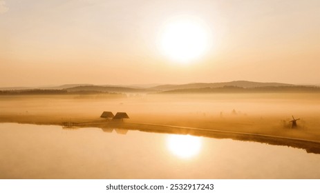 A misty sunrise illuminates a serene landscape with distant hills, a winding road by calm water, and a quaint windmill beside rustic houses. - Powered by Shutterstock