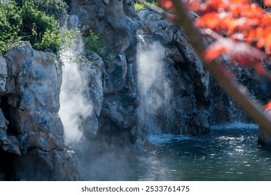 Misty waterfalls cascade down rocky cliffs into a serene pond, surrounded by lush greenery and highlighted by vibrant red leaves. - Powered by Shutterstock