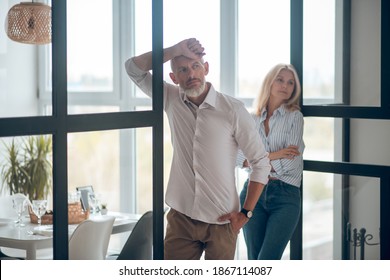 Misunderstanding. Bearded Man Looking Sad, Blonde Woman Watching Him With Arms Crossed