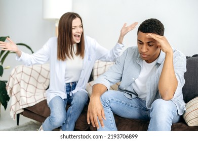 Misunderstanding In Relationships, Jealousy, Quarrel. A Married Multiracial Couple Sorts Things Out While Sitting In The Living Room On A Sofa, A Caucasian Woman Screams At Her Hispanic Husband