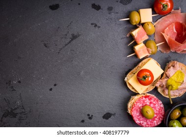 Mix Of Different Snacks And Appetizers. Spanish Tapas On A Black Stone Background. Tapas Bar. Space For Text. Deli, Sandwiches, Olives, Sausage, Anchovies, Cheese, Jamon, Pepper, Tomatoes. Top View