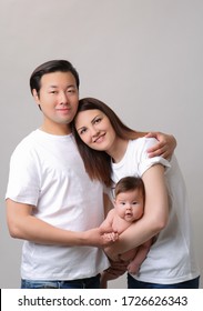 Mix Face Family With Baby. Chinese, European. Parents Are Holding Baby In Their Arms. Smiling, Hugging, Happy. Studio Photo. White Background. Clothing Casual. Concept Of Traditions, Unification, Love