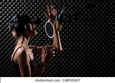 Mix Race Skinny Woman Blonde Hair Cap Earmuff Headphone Sing A Song Loudly With Power Sound Over Hanging Microphone Condenser. Egg Crate Studio Low Lighting Shadow Sound Proof Absorbing Wall Room