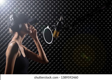 Mix Race Skinny Woman Blonde Hair Cap Earmuff Headphone Sing A Song Loudly With Power Sound Over Hanging Microphone Condenser. Egg Crate Studio Low Lighting Shadow Sound Proof Absorbing Wall Room