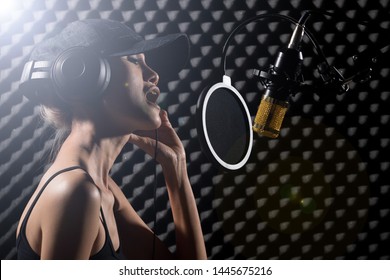 Mix Race Skinny Woman Blonde Hair Cap Earmuff Headphone Sing A Song Loudly With Power Sound Over Hanging Microphone Condenser. Egg Crate Studio Low Lighting Shadow Sound Proof Absorbing Wall Room