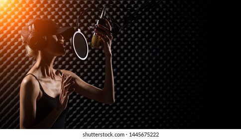 Mix Race Skinny Woman Blonde Hair Cap Earmuff Headphone Sing A Song Loudly With Power Sound Over Hanging Microphone Condenser. Egg Crate Studio Low Lighting Shadow Sound Proof Absorbing Wall Room