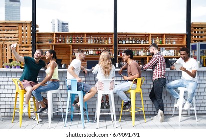 Mix-Rennteam aus stylischen, niedlichen und kreativen jungen Leuten Kollegen arbeiten und entspannen sich gemeinsam auf kreativem Arbeitsbereich auf der offenen Terrasse hinter einer weißen Theke in der Nähe von Holzstühlen mit Getränken