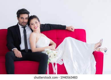 Mix Race Wedding Concept, Beautiful Asian Woman Lie Down On Chest Of Caucasian Handsome Man On Red Sofa Bed, Happy Couple Spent Time Together, Man Taking Care Bride With Love And Tenderness