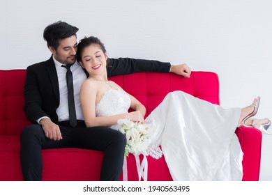 Mix Race Wedding Concept, Beautiful Asian Woman Lie Down On Chest Of Caucasian Handsome Man On Red Sofa Bed, Happy Couple Spent Time Together, Man Taking Care Bride With Love And Tenderness